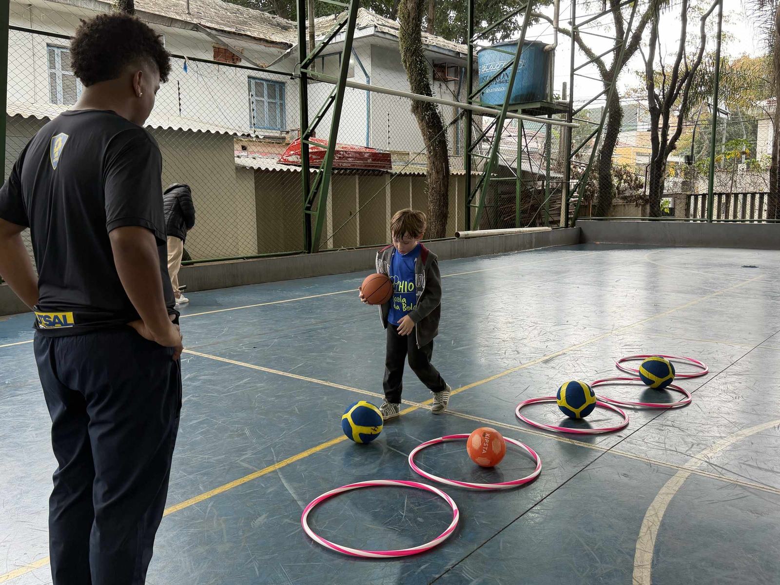 Menino do H10 Kids com a bola de basquete no circuito de arcos, com o professor por perto
