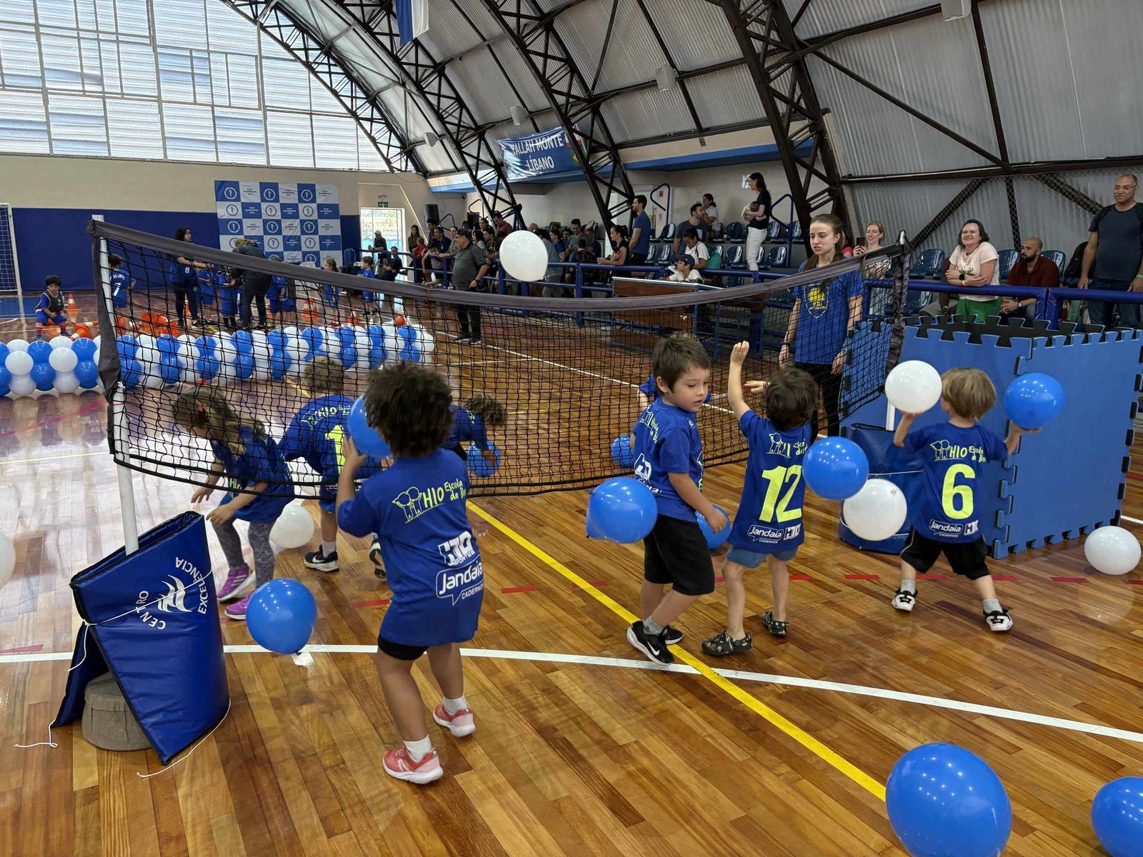 Turma do H10 Kids brincando com bexigas em volta da rede, dentro do ginásio